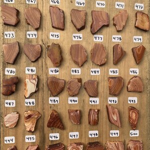 K&ouml;nnte beinhalten: Eine Sammlung von 25 kleinen, unregelm&auml;&szlig;ig geformten, polierten Steinen mit roten, orangen und wei&szlig;en B&auml;ndern. Die Steine sind in Reihen auf einer Holzoberfl&auml;che angeordnet und von 466 bis 500 nummeriert.