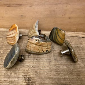 K&ouml;nnte beinhalten: Eine Sammlung dekorativer Schrankkn&ouml;pfe mit Naturstein-Designs. Die Kn&ouml;pfe weisen verschiedene Formen und Farben auf, darunter Beige, Grau und Gelb, mit Metallbeschl&auml;gen zur Befestigung. Die Kn&ouml;pfe sind auf einer Holzoberfl&auml;che angeordnet.