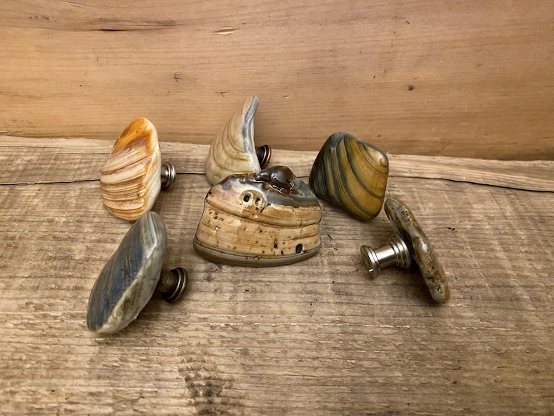 K&ouml;nnte beinhalten: Eine Sammlung dekorativer Schrankkn&ouml;pfe mit Naturstein-Designs. Die Kn&ouml;pfe weisen verschiedene Formen und Farben auf, darunter Beige, Grau und Gelb, mit Metallbeschl&auml;gen zur Befestigung. Die Kn&ouml;pfe sind auf einer Holzoberfl&auml;che angeordnet.