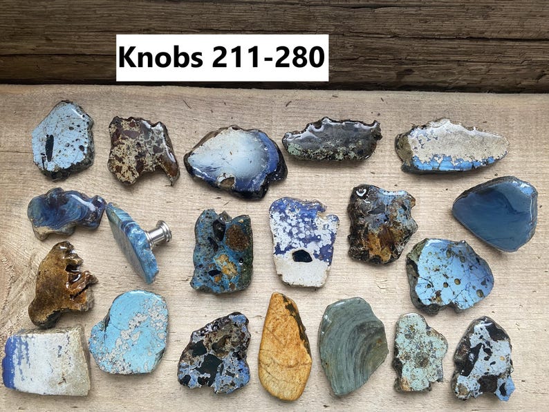 May include: A collection of 17 ceramic knobs in various shades of blue, brown, and white. The knobs are irregularly shaped and have a rustic, handmade appearance. The text "Knobs 211-280" is visible at the top of the image.