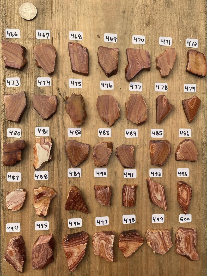 Op de afbeelding: Een verzameling van 25 kleine, onregelmatig gevormde, gepolijste stenen met rode, oranje en witte banden. De stenen zijn gerangschikt in rijen op een houten oppervlak en genummerd van 466 tot 500.