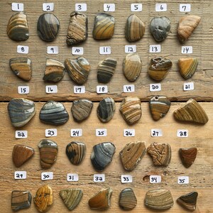 K&ouml;nnte beinhalten: Eine Sammlung von f&uuml;nfunddrei&szlig;ig polierten Steinen mit verschiedenen Farben und Mustern, darunter Streifen und Wirbel. Die Steine sind auf einer Holzoberfl&auml;che angeordnet, jede mit einer Nummer von 1 bis 35 versehen. Die Steine variieren in Gr&ouml;&szlig;e und Form, mit Farben von Braun bis Grau.