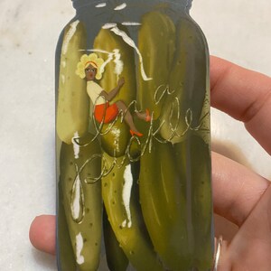 May include: A sticker of a glass jar filled with green pickles. A woman with blonde hair and an orange dress is climbing up the side of the jar. The text "In a pickle" is written on the jar.