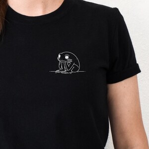 May include: Black t-shirt featuring a small white outline drawing of a dejected frog on the upper left chest. The shirt has short sleeves and is made of a soft material. The person is wearing blue jeans.