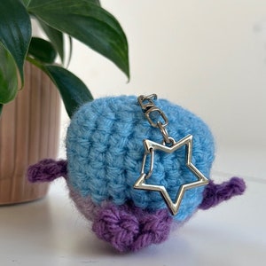 May include: A handmade crochet keychain in the shape of a bird. The bird is blue and purple with a silver star charm attached. The keychain is on a white surface with a green plant in the background.