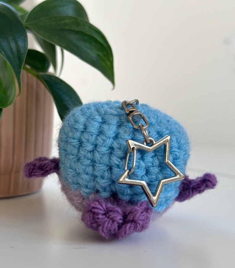 May include: A handmade crochet keychain in the shape of a bird. The bird is blue and purple with a silver star charm attached. The keychain is on a white surface with a green plant in the background.
