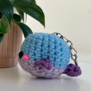 May include: A handmade crochet whale keychain in shades of blue and purple. The whale has a black eye, pink cheeks, and a small purple tail. A silver-colored metal clasp is attached to the top.