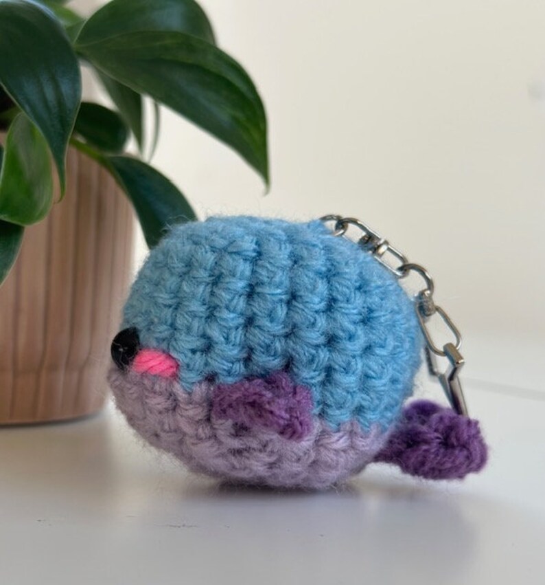 May include: A handmade crochet whale keychain in shades of blue and purple. The whale has a black eye, pink cheeks, and a small purple tail. A silver-colored metal clasp is attached to the top.