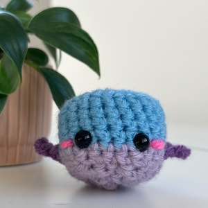 May include: A handmade crochet whale in shades of blue and lavender. The whale has black safety eyes, pink cheeks, and small purple fins. The amigurumi is sitting on a white surface.