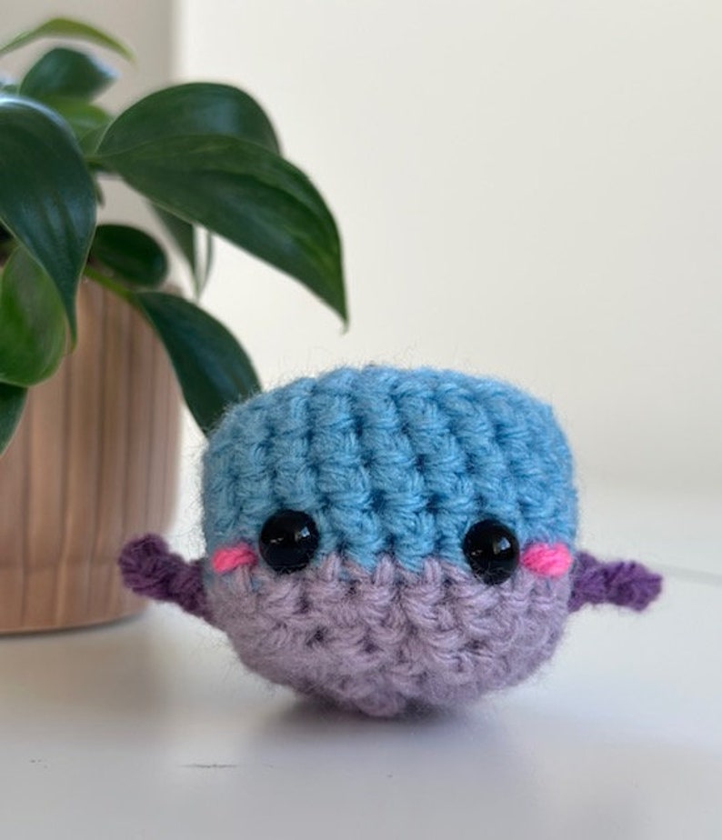 May include: A handmade crochet whale in shades of blue and lavender. The whale has black safety eyes, pink cheeks, and small purple fins. The amigurumi is sitting on a white surface.