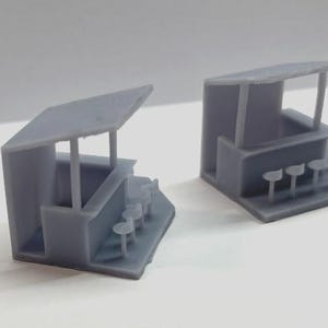 Può includere: Due edifici in miniatura di colore grigio con tetto piatto e bancone. Ogni edificio ha un piccolo baldacchino e una fila di sedie in miniatura davanti al bancone.