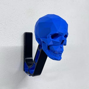 Puede incluir: Un colgador de pared con un diseño de calavera geométrica azul brillante. La calavera está montada sobre un soporte negro y azul, creando un contraste llamativo contra la pared blanca. Este gancho único añade un toque de decoración atrevida.