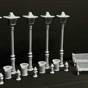 Op de afbeelding: Een verzameling miniatuur grijze straatlantaarns, vuilnisbakken, brandkranen en banken. De lampen hebben een klassiek ontwerp met een vierkante bovenkant. De scène speelt zich af tegen een donkere achtergrond.