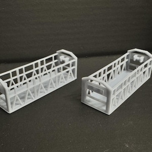 Puede incluir: Dos estructuras de color gris claro, con forma de jaula, de forma rectangular, con soportes angulados y una característica similar a un asa en cada extremo. Los objetos están sobre un fondo oscuro.