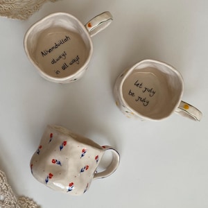 Pode incluir: Três canecas de cerâmica com padrões florais. Uma caneca tem o texto "Alhamdulillah sempre em todos os momentos" escrito dentro. Outra caneca tem o texto "que julho seja julho" escrito dentro. A terceira caneca tem um padrão floral vermelho e azul.