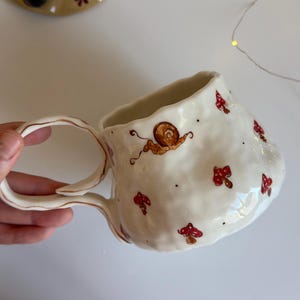 Könnte beinhalten: Ein cremefarbener Keramikbecher mit unebener, strukturierter Oberfläche. Der Becher ist mit handgemalten roten Pilzen und einer braunen Schnecke verziert, akzentuiert durch kleine schwarze Punkte. Er hat einen geschwungenen, welligen Henkel.
