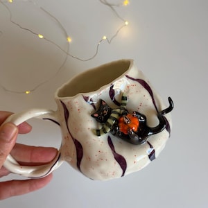 Può includere: Una tazza in ceramica bianca con bordo e manico ondulati e irregolari, caratterizzata da strisce verticali viola e macchie rosse. Un gatto nero scolpito con una sciarpa a righe verdi e bianche è applicato sul lato e tiene in mano una piccola zucca arancione.