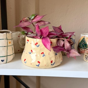 Può includere: Un piccolo vaso in ceramica color crema con un motivo floreale di fiori rossi e dettagli blu. Il vaso contiene una vivace pianta viola con foglie lunghe e appuntite. Altri pezzi decorativi in ceramica sono visibili sullo scaffale.