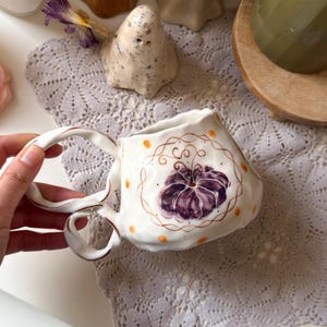 Könnte beinhalten: Ein weißer Keramikbecher mit gewelltem, unregelmäßigem Rand und einem Doppelhenkel. Der Becher ist mit einem handgemalten violetten Kürbismotiv verziert, das von einer braunen Kreislinie umrahmt und mit kleinen orangefarbenen Punkten versehen ist.