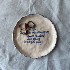 May include: A white ceramic trinket dish with a blue rim and blue handwritten text. The dish holds three rings. The text reads: "if life is going to be exhausting anyway I choose to be tired while chasing beautiful things."