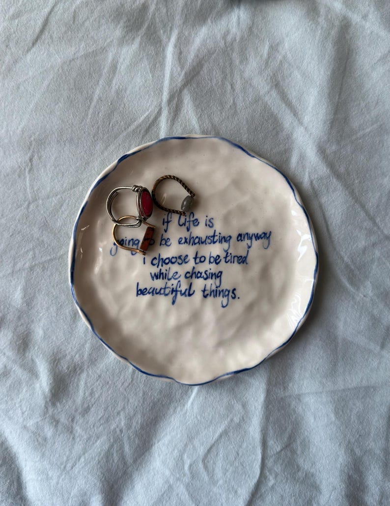 May include: A white ceramic trinket dish with a blue rim and blue handwritten text. The dish holds three rings. The text reads: "if life is going to be exhausting anyway I choose to be tired while chasing beautiful things."