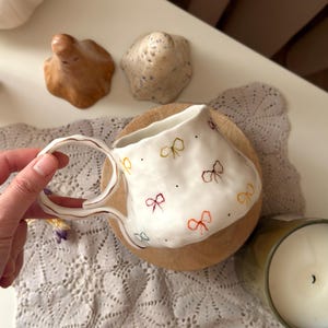 Peut inclure: Une tasse en céramique blanche avec un bord irrégulier et ondulé ainsi qu'une surface texturée. La tasse est décorée de petits nœuds peints à la main de différentes couleurs, notamment jaune, marron, rouge et bleu, parsemés de points noirs.