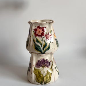 Puede incluir: Un recipiente de cerámica con borde festoneado y acabado brillante en color blanco roto, con motivos florales pintados a mano en tonos rojo, morado y verde. La superficie está decorada con delicadas líneas y puntos de color marrón oscuro.