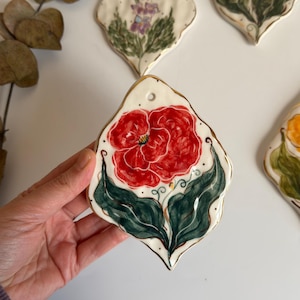 May include: A leaf-shaped ceramic ornament featuring a hand-painted red flower with dark green leaves and gold-painted edges. The piece has an irregular, organic outline and a small circular hole at the top for hanging, showcasing a glossy finish.