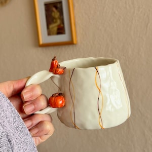 Peut inclure: Une tasse en céramique de couleur crème avec une surface texturée et irrégulière, dotée d'une anse ornée de trois petites citrouilles sculptées orange. La tasse présente de fines lignes verticales ondulées brunes et orange sur l'extérieur.