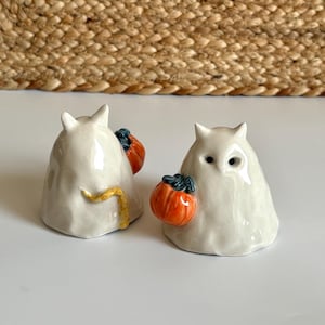 May include: Two white ceramic ghost figurines, each featuring pointed ears and holding a small orange pumpkin with a dark blue stem. One ghost is shown from the front with visible eye holes, while the other is viewed from the side with a yellow tail.