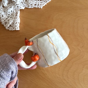 Può includere: Tazza in ceramica bianca con linee arancioni e marroni e due piccole decorazioni a forma di zucca arancione sul manico. La tazza ha una forma irregolare unica, perfetta per l'autunno. È un articolo fatto a mano.