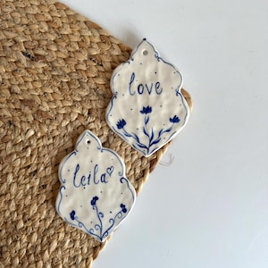 May include: Two white ceramic wall hangings with a blue floral design and the words "love" and "Leila" written in blue script. The hangings have a decorative shape and a small hole for hanging.