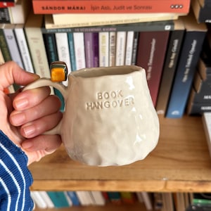 May include: Cream-colored ceramic mug with the text "BOOK HANGOVER" embossed on the front. The mug has a textured surface and a rounded shape. A ring with a square, orange stone is on the handle. Bookshelves are in the background.
