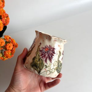 Könnte beinhalten: Ein cremefarbenes Keramikgefäß mit einem ausgestellten, gewellten Rand, verziert mit handgemalten Illustrationen von dunkelvioletten Blumen mit orangefarbenen Mitten und grünen Ranken, akzentuiert durch kleine rote Sprenkel.