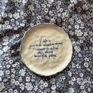 May include: A round, off-white ceramic plate with a wavy edge and a blue rim. The plate features the handwritten quote, "if life is going to be exhausting anyway I choose to be tired while chasing beautiful things."