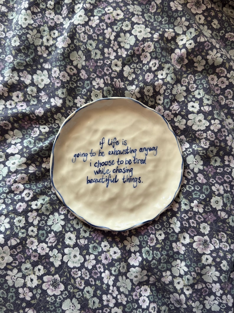 May include: A round, off-white ceramic plate with a wavy edge and a blue rim. The plate features the handwritten quote, "if life is going to be exhausting anyway I choose to be tired while chasing beautiful things."