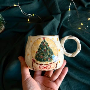 May include: A ceramic mug with a hand-painted Christmas tree design. The mug has a cream-coloured base with a round image of a green Christmas tree decorated with red bows and a star on top. The handle is a twisted rope design.