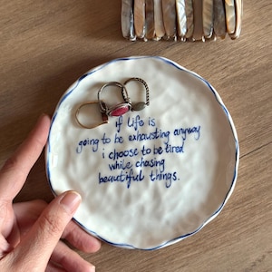 May include: A white ceramic trinket dish with a blue rim and handwritten text. The text reads "if life is going to be exhausting anyway I choose to be tired while chasing beautiful things." Two rings rest on the dish, and a beaded bracelet is above.