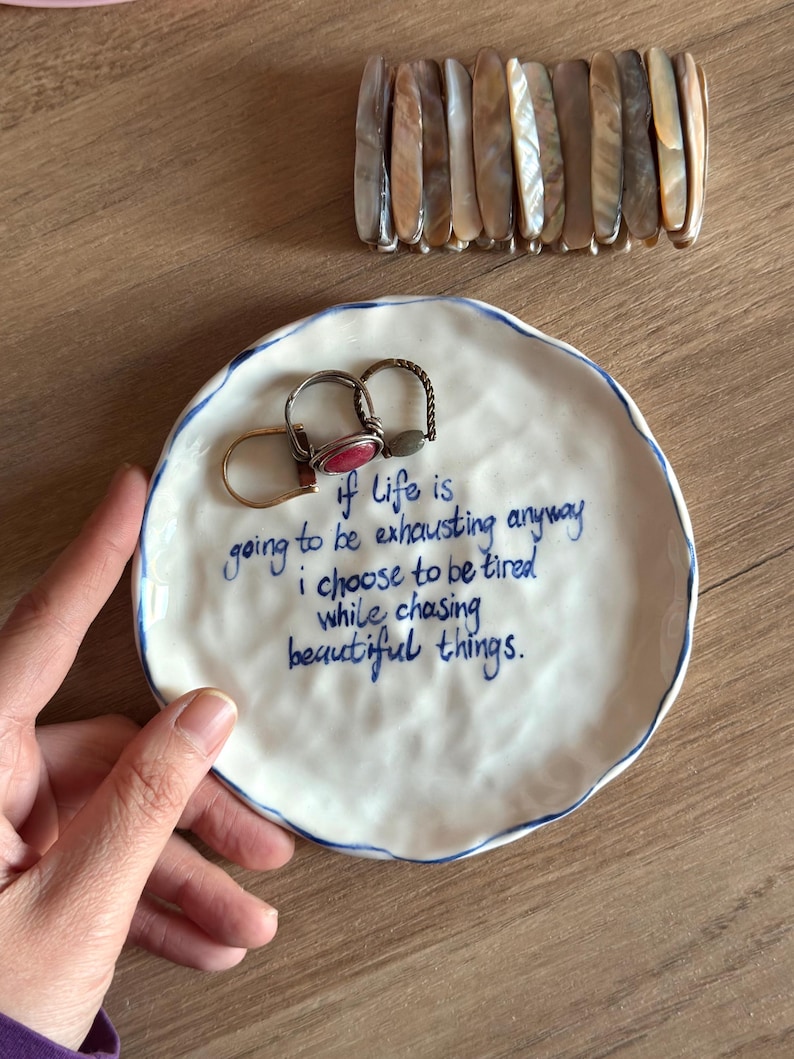 May include: A white ceramic trinket dish with a blue rim and handwritten text. The text reads "if life is going to be exhausting anyway I choose to be tired while chasing beautiful things." Two rings rest on the dish, and a beaded bracelet is above.