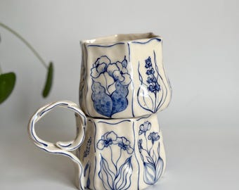 Puede incluir: Dos tazas de cerámica apiladas con base color crema y diseños florales azules. Las tazas tienen una forma cuadrada única, con un asa en la taza inferior. Los patrones florales incluyen varias flores y hojas.