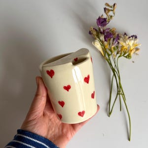 Può includere: Una tazza in ceramica color crema decorata con cuori rossi. La tazza ha un design unico del manico ed è tenuta in mano da una persona. Fiori secchi sullo sfondo.