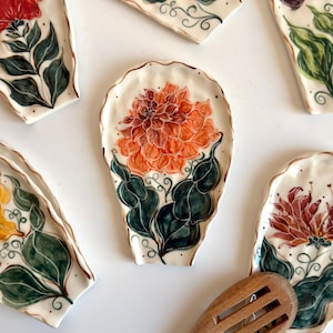May include: A collection of five ceramic spoon rests with irregular, scalloped edges. Each piece features hand-painted floral designs in shades of orange, red, and yellow, paired with dark green leaves and swirling vine details on a white background.