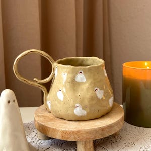 Peut inclure: Une tasse en céramique jaune clair mouchetée avec une surface ondulée et irrégulière et une grande anse dorée de forme irrégulière. La tasse est décorée de petits fantômes blancs peints à la main avec des visages simples sur l'extérieur.