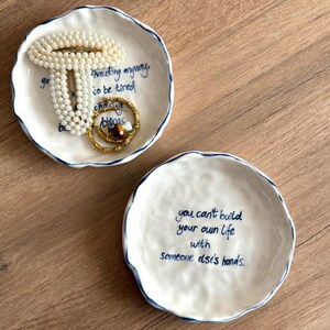 May include: Two white ceramic trinket dishes with blue rims and handwritten text. One dish holds a pearl bracelet and a gold ring. The other dish has the text: "you can't build your own life with someone else's hands."