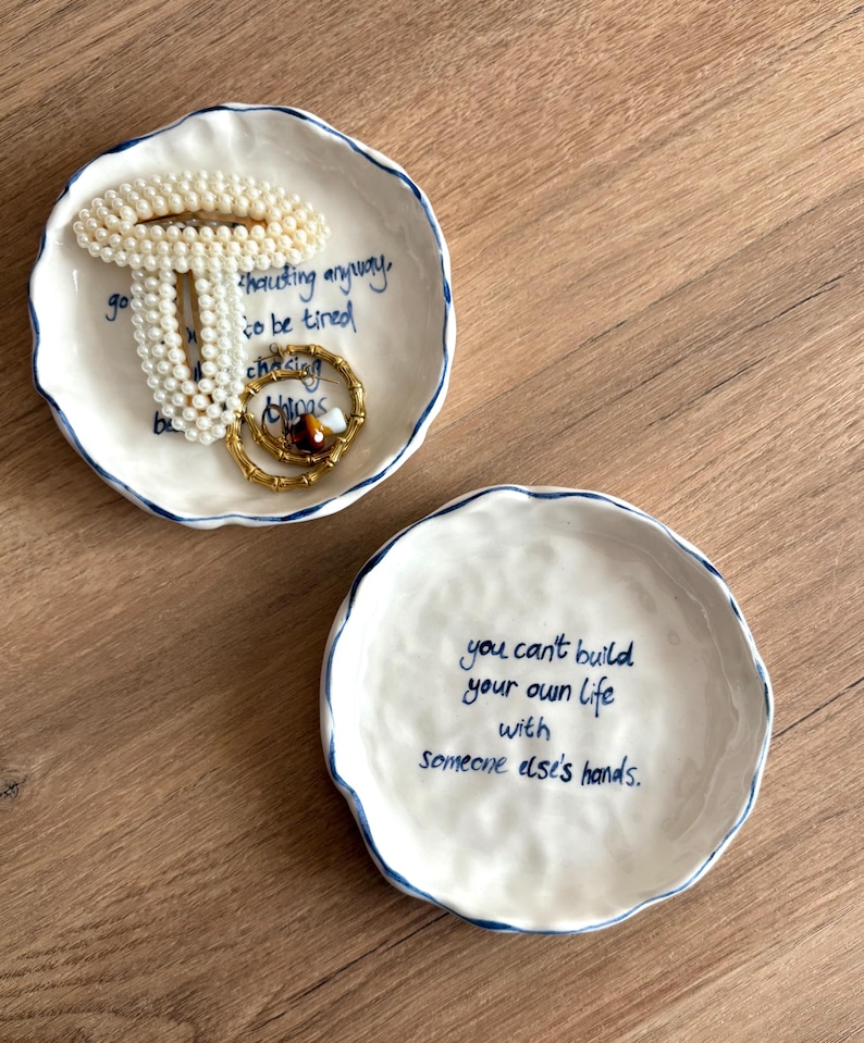 May include: Two white ceramic trinket dishes with blue rims and handwritten text. One dish holds a pearl bracelet and a gold ring. The other dish has the text: "you can't build your own life with someone else's hands."