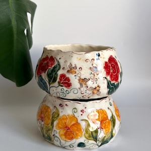 May include: Two stacked ceramic bowls with a cream-colored glaze and scalloped edges. The top bowl features hand-painted red flowers and small bees. The bottom bowl displays hand-painted yellow flowers with green leaves and swirling vine details.
