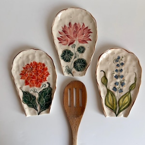 May include: Three ceramic spoon rests with floral designs. The spoon rests are off-white with brown edges and feature hand-painted flowers in shades of orange, pink, and blue. A wooden slotted spoon rests between the spoon rests.