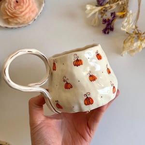 Puede incluir: Una taza de cerámica de color crema con una forma irregular hecha a mano y un asa grande y curva. La superficie exterior presenta un patrón repetitivo de pequeñas calabazas naranjas pintadas a mano con tallos marrones y puntos negros.