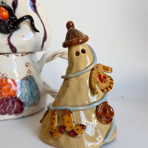 Op de afbeelding: Een beige keramieken spookfiguurtje met een gestreepte muts, een blauwe sjaal en versierd met kleine gebeeldhouwde items zoals een want, een hoed en een paar sokken. Daarachter staat een witte keramieken kan met geschilderde pompoenen.