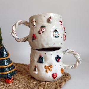 Puede incluir: Dos tazas de cerámica blanca apiladas, decoradas con adornos navideños. Cada taza tiene un asa retorcida con detalles en rojo y verde. Las tazas están adornadas con miniaturas.